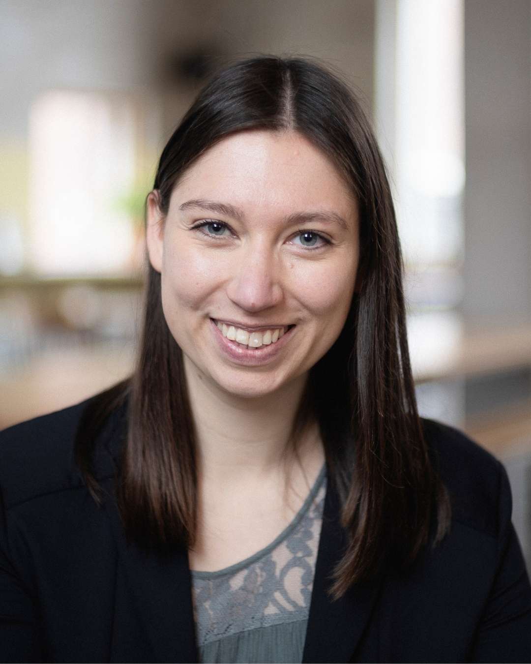 Porträtfoto einer lächelnden Frau mit langen dunklen Haaren in schwarzem Blazer vor unscharfem Innenraum-Hintergrund.