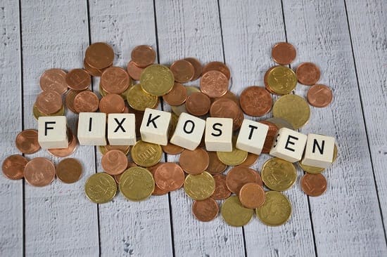 Holzblöcke bilden das Wort "Fixkosten" mit Münzen auf dem Hintergrund